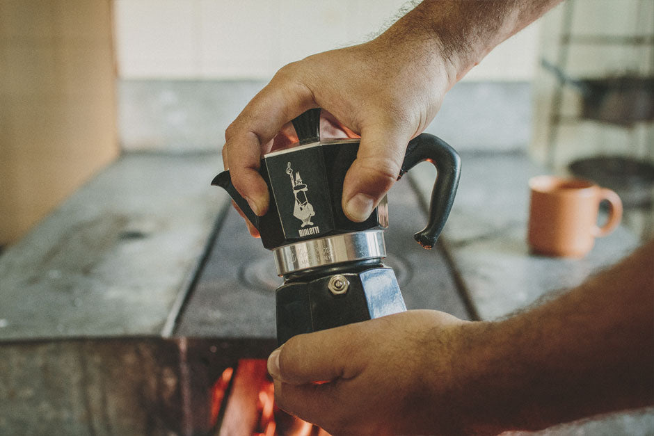 Homem segurando uma cafeteira Moka preta em frente a fogão de lenha