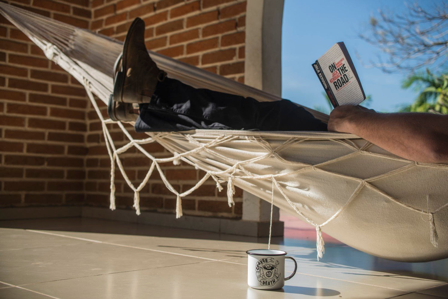 homem deitado na rede lendo livro com caneca em baixo