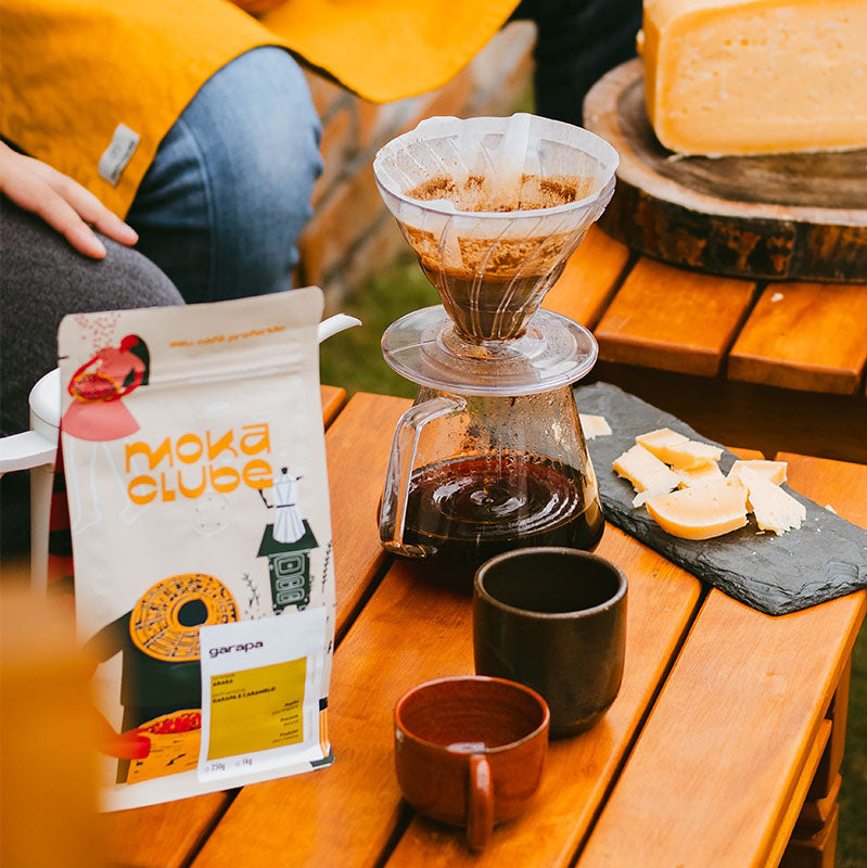 Uma mesa com pacote de café garapa xicaras, queijo e um café sendo coado