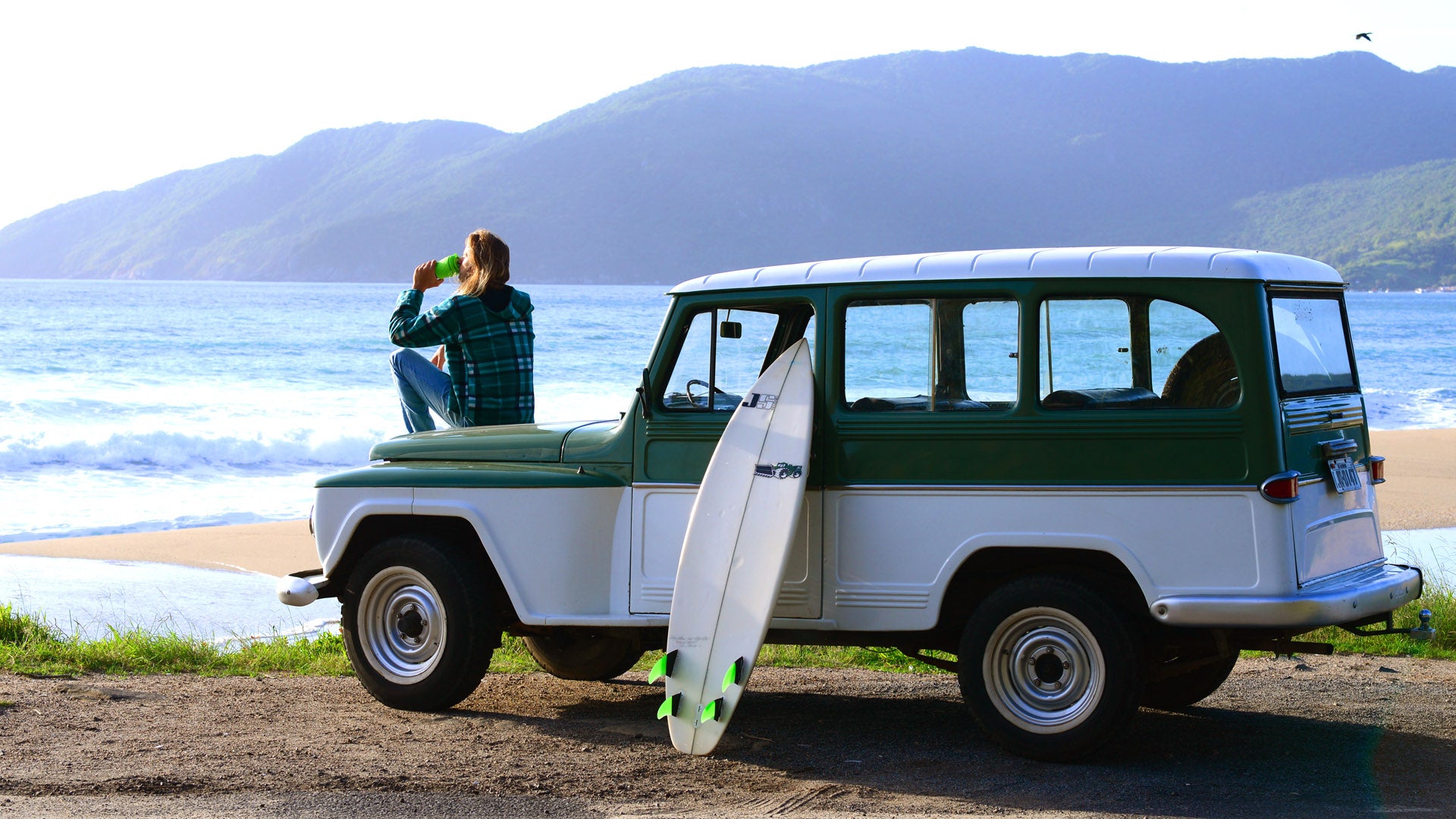 Homem bebendo em uma pressca verde de frente para o mar sentado em frente ao seu carro