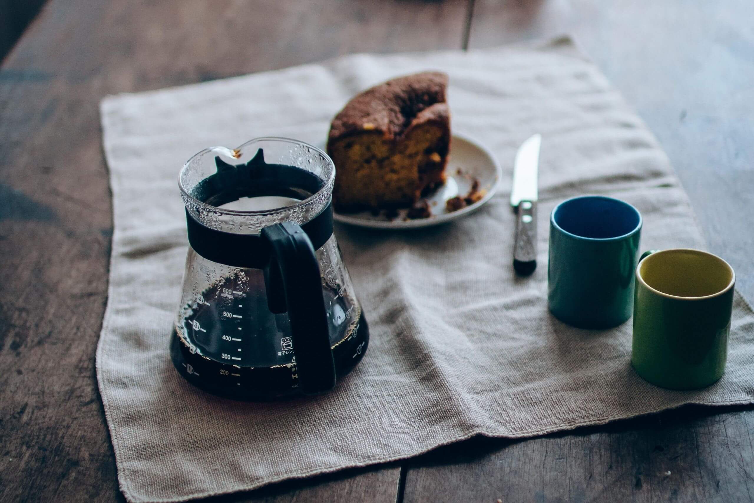 Mesa com um pedaço de bolo de chocolate, com uma xícara de café quase vazia e duas canecas coloridas.