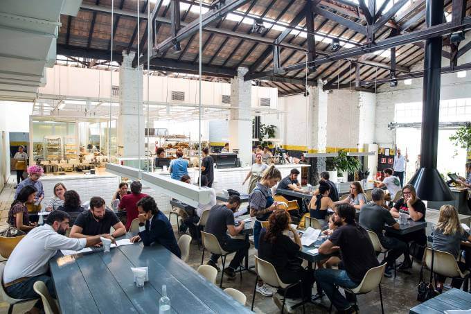 Cafeteria Futuro Refeitório em horário de pico, com mesas de madeira e cadeiras de plástico ocupadas por diversas pessoas.